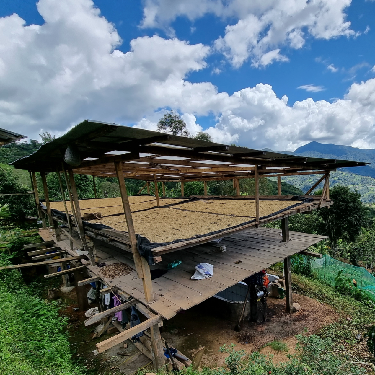 A rustic wooden elevated structure with a partially collapsed or missing roof sits on a hillside in a mountainous rural setting under a blue sky with large white clouds. Green vegetation and distant hills are visible.