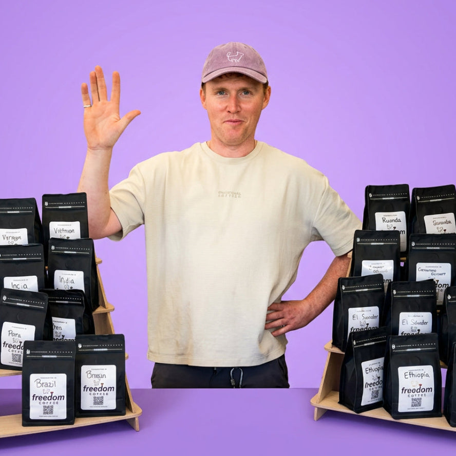 A man wearing a beige t-shirt and a purple cap stands behind a table displaying many bags of coffee under a white tent. The table has a purple banner that reads 'freedom COFFEE'.